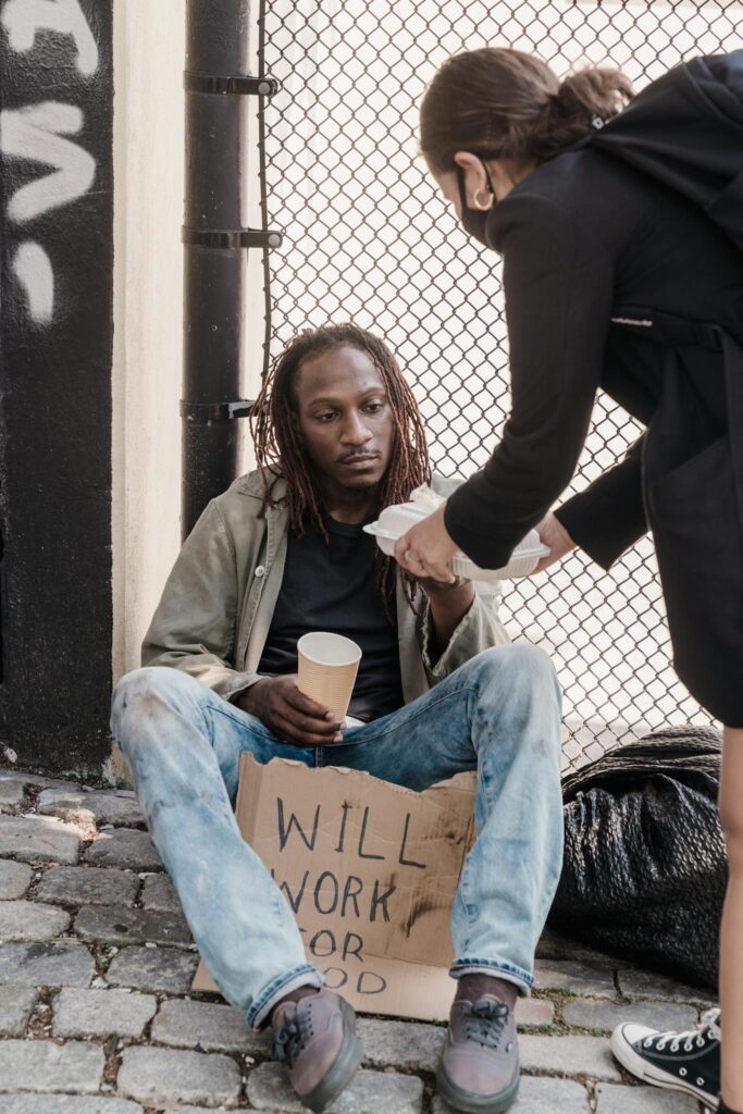 A woman offers food to a man sitting on the sidewalk holding a sign, showcasing empathy.